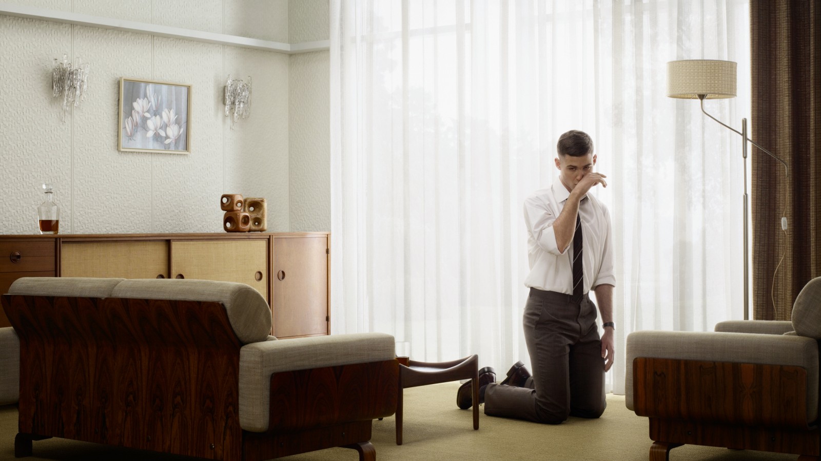 A man is kneeling in the floor, crying. He's a young man dressed with a formal shirt and trousers, wearing a tie. He looks somehow angry and sad, and he's looking at some point out of camera, to the left of the scene. The room is light, empty, looks like a 60's decorated scene in the USA. It's very plain, there are furniture and decorations, elegant, but doesn't look like a home, more like an hotel.